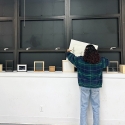 A student is standing in a room, facing away from the camera, organizing or examining items on a counter beneath large wall-mounted cabinets. The cabinets are open, displaying empty shelves. Atop the counter are various books and small boxed items.
