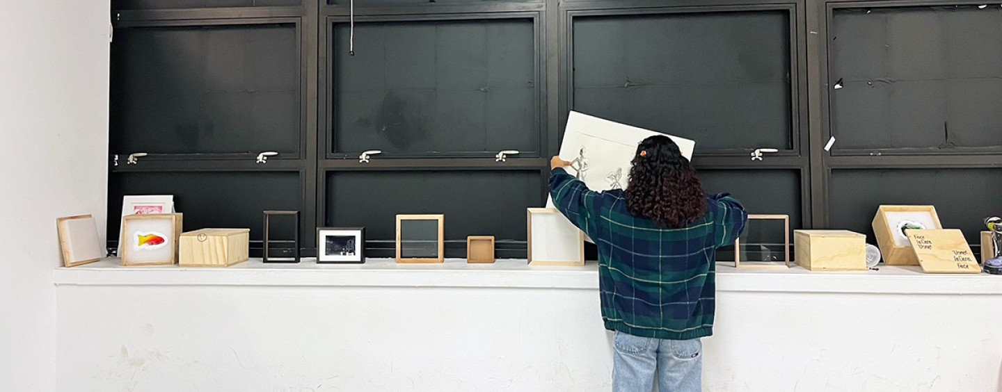 A student is standing in a room, facing away from the camera, organizing or examining items on a counter beneath large wall-mounted cabinets. The cabinets are open, displaying empty shelves. Atop the counter are various books and small boxed items.